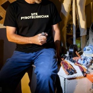 A handsome Foxfur wearing a custom Foxfur Ink black t-shirt with white lettering that says site pyrotechnician. The scene is nighttime at burning man, he is sitting beside an effigy with road flares that have electric matches duct taped to the ignition compound getting ready to set the effigy on fire.